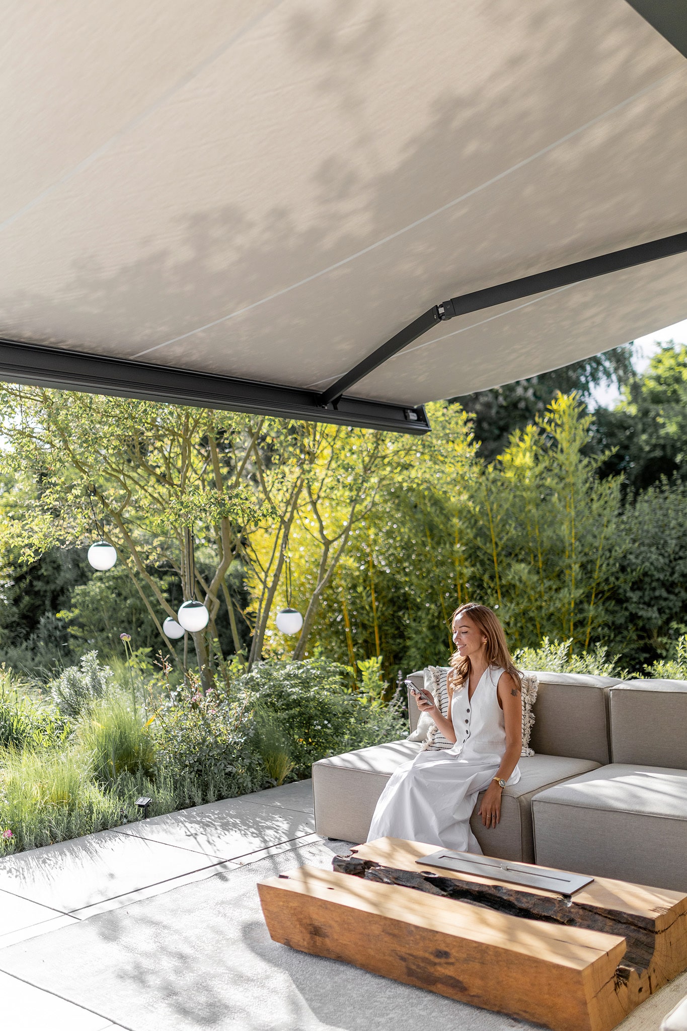 Ein Terrassenbereich mit beiger Markise in dunkelgrauem Rahmen, Outdoor‑Couch auf der eine Frau im Schatten entspannt von Warema.