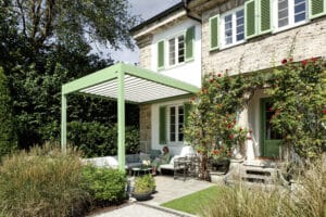 Klassische Villa mit verspieltem Garten und einem Außenbereich mit grüner Pergola - passend zu den Fensterläden - und einem halboffenen Lamellendach über einer Terrasse mit Sitzgruppe von Warema.