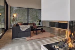 Ein moderner Wohnraum mit Kamin, Eckverglasung und Terrassenschiebetüren mit schwarzem Rahmen und passenden Aussenjalousien schafft elegante harmonische Lichtstimmung von WAREMA.