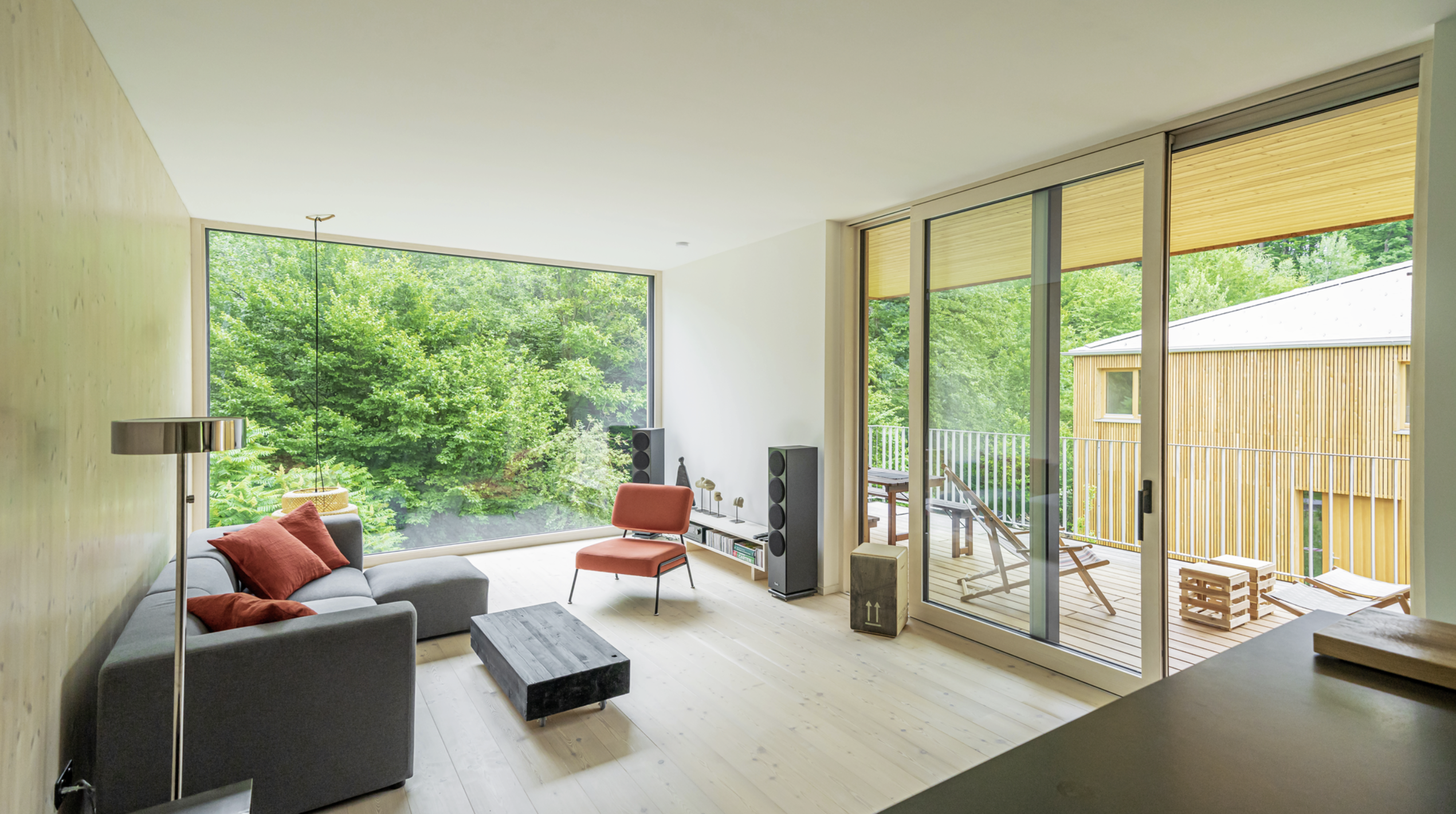Weissenseer Holz-System-Bau zeigt lichtdurchflutetes Wohnzimmer mit Wandvertäfelung, bodentiefer vollverglasten Front und Balkonschiebtür in einem Holzriegelhaus.