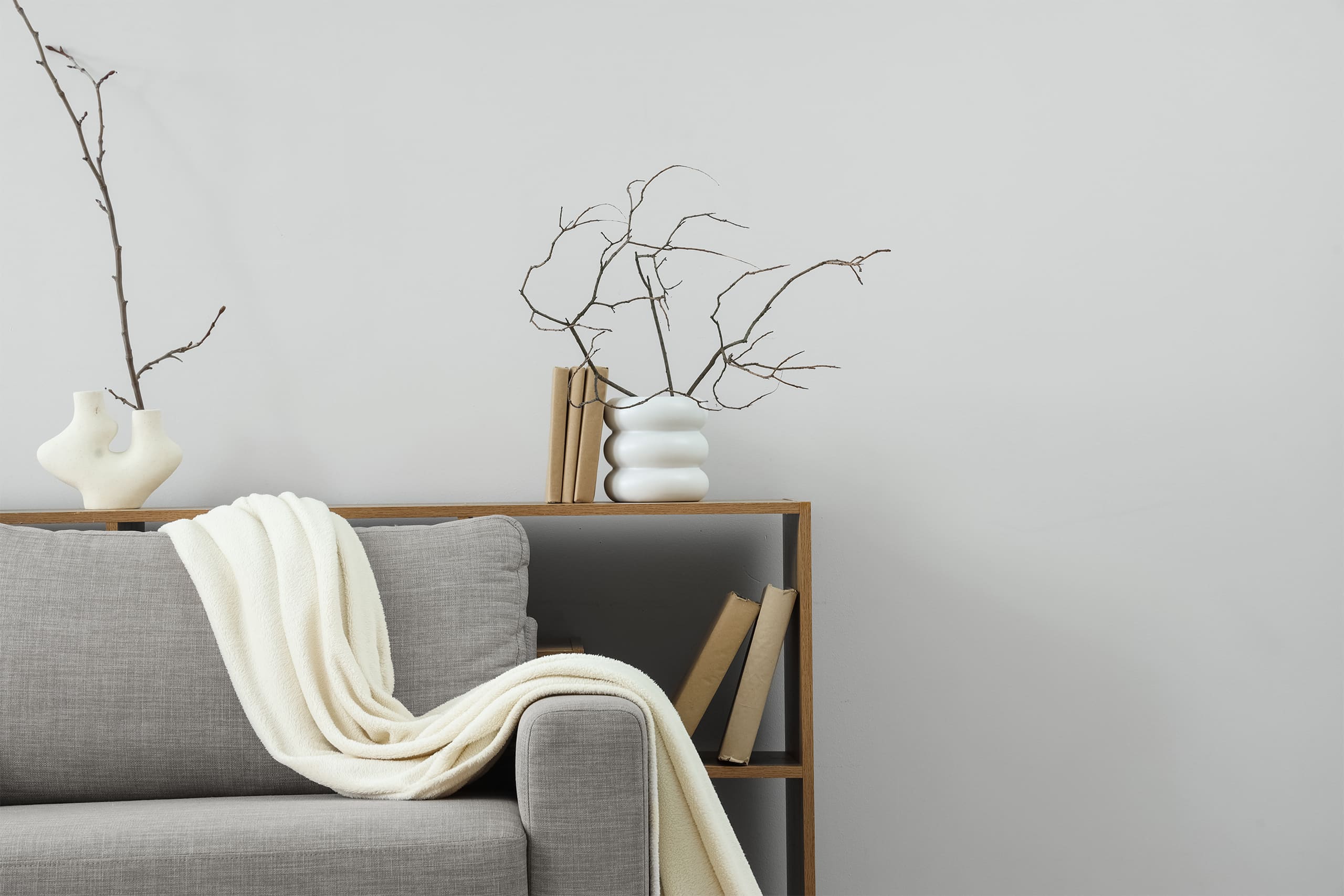 Bücher und Vasen mit getrockneten Zweigen in einem Regal aus hellem Holz im Wohnzimmer. Davor steht ein bequemes Sofa in Grau, mit einer weißen Tagesdecke trappiert. - KI-generiert