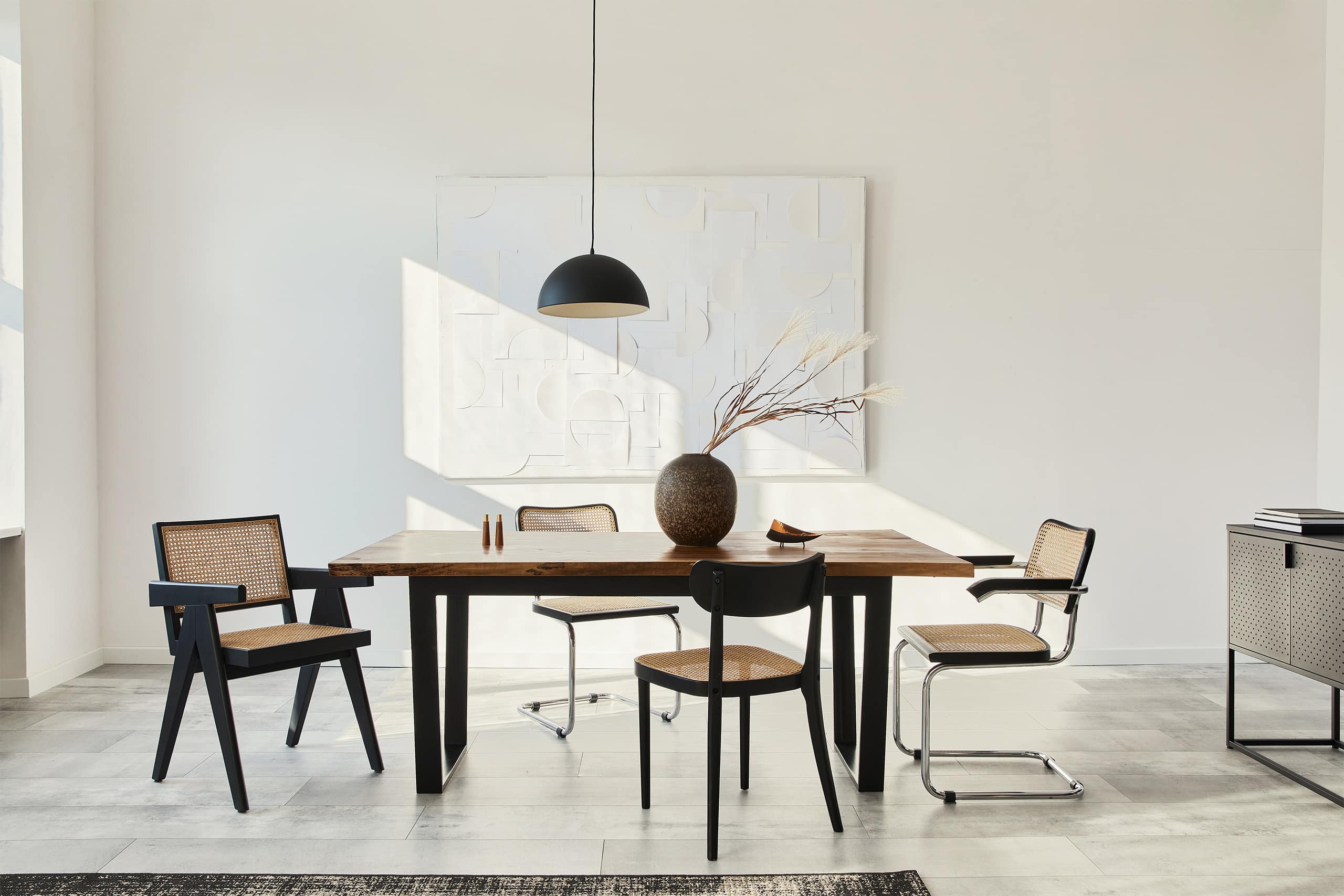 Minimalistische Komposition eines Esszimmerinterieurs bzw. Esszimmer / Essbereich mit Holztisch, Designstühlen, Trockenblumen in einer Vase, schwarzer Pendelleuchte, Kunstwerken an der Wand und eleganten persönlichen Accessoires.