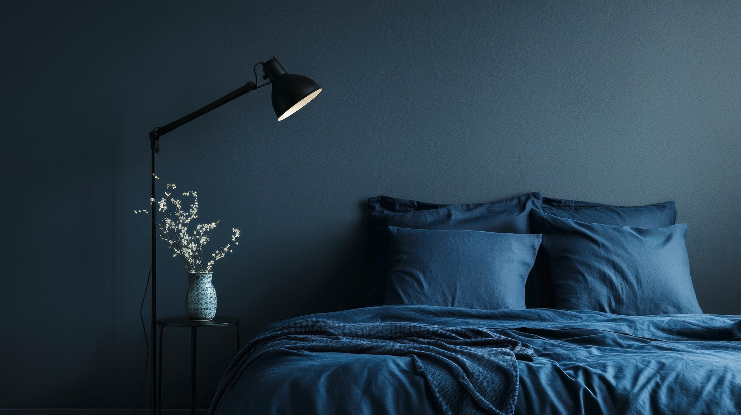Elegenates Schlafzimmer mit dunkelblauer Wandfarbe und Satin-Bettwäsche in dazu passendem Royalblau. ein keiner, schwarzer Nachttisch mit einer schwarzen Tischleuchte ergänzen den edlen Look. - KI-generiert
