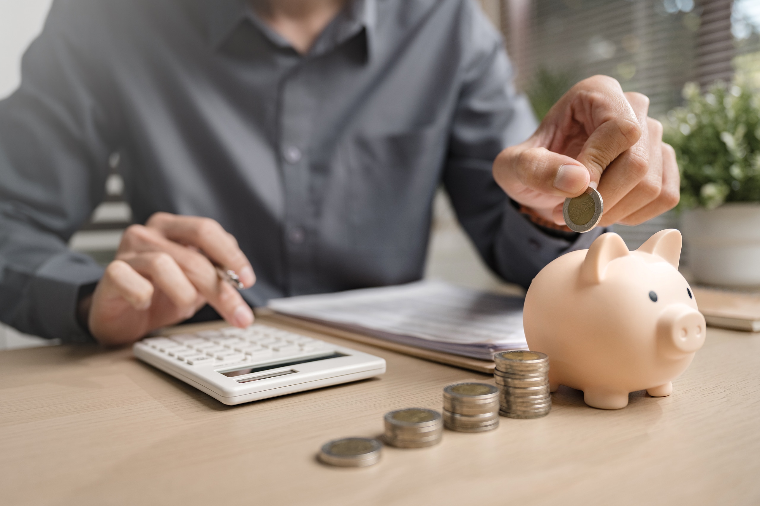 Mann erstellt gerade einen finanziellen Kalkulation und plant seine zukünftigen Ausgaben mithilfe eines Taschenrechners und wirft Münzen in ein Sparschwein.