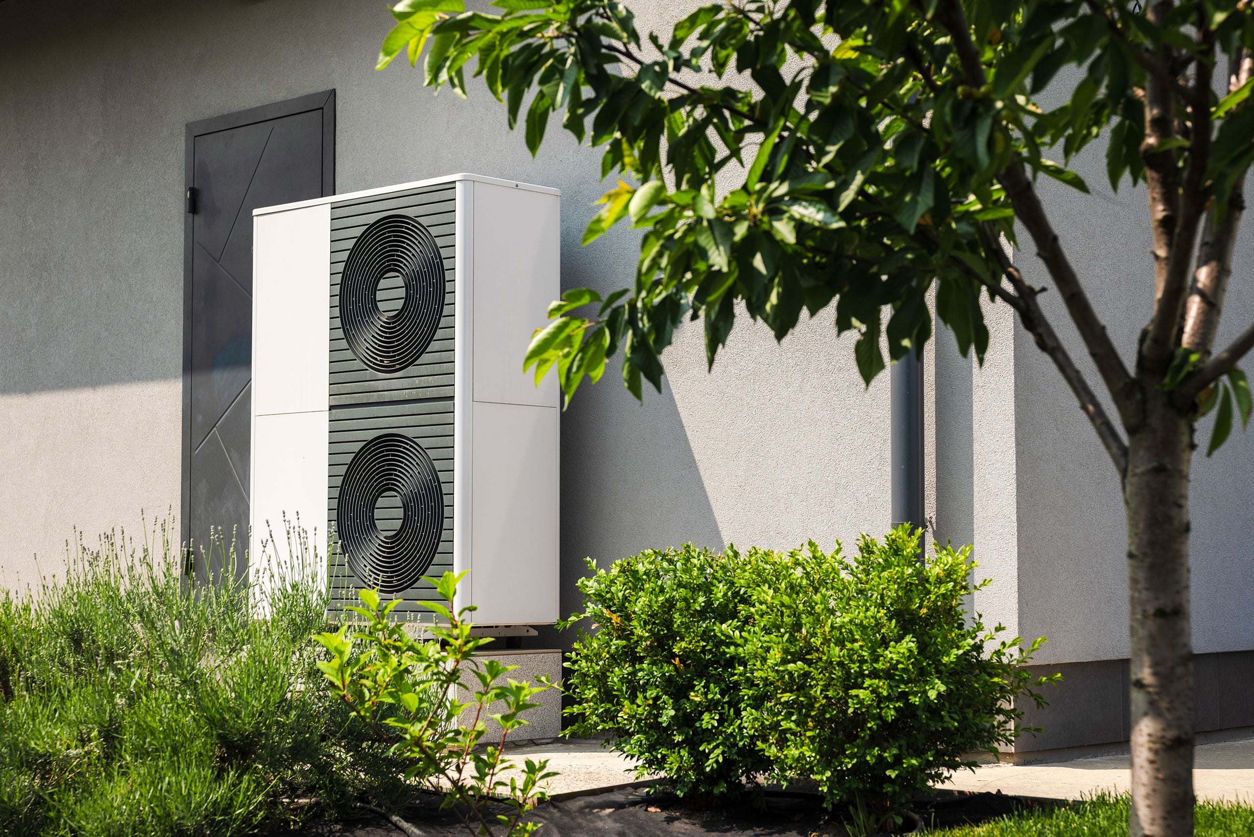 Moderne Außenwärmepumpe, installiert in der Nähe einer Hauswand, umgeben von grünen Sträuchern und einem Baum im angelegten Garten.