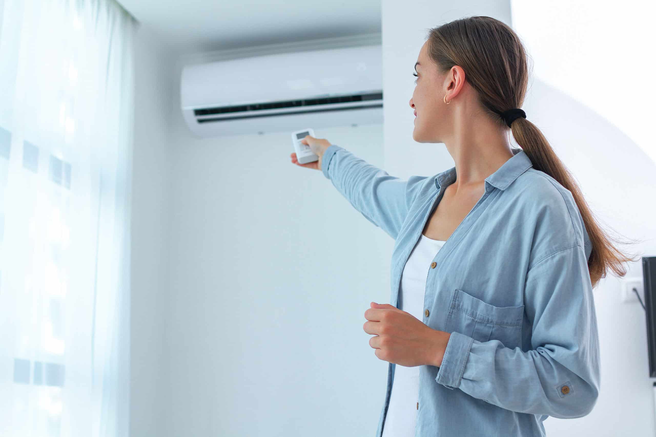 Eine junge Frau reguliert die Temperatur der Klimaanlage in ihrem Zimmer zu Hause mithilfe der Fernbedienung.