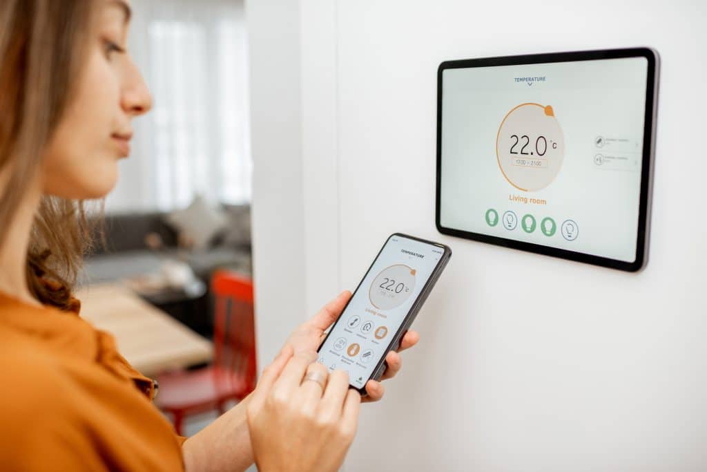 Junge Frau regelt die Temperatur im Wohnzimmer mit Smartphone und digitalem Touchscreen. Konzept der Heizungssteuerung im Smart Home.