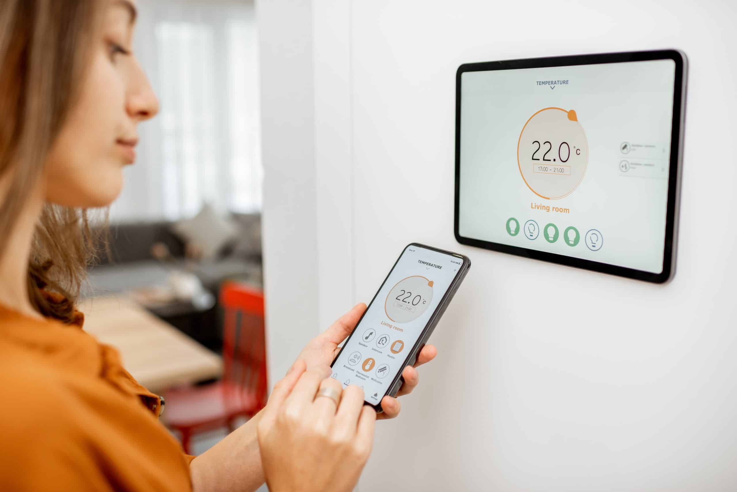 Junge Frau regelt die Temperatur im Wohnzimmer mit Smartphone und digitalem Touchscreen. Konzept der Heizungssteuerung im Smart Home.